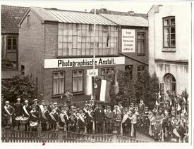 1923 Vor der Photographischen Anstalt von Ludwig Behning am Markt ist die Musikgruppe (Trommler und Pfeifer Corps) der Kinder-Schützengilde mit Flaggen, Standarten und dem hölzernen Adler sowie einer aus Nadelgehölz gebundenen Krone angetreten